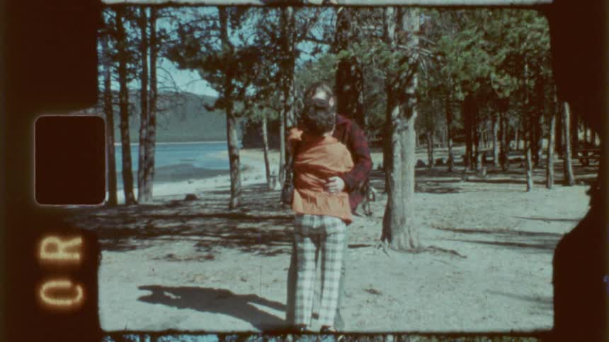 A man kisses a woman in a forest near a lake while making a peace sign with his hand