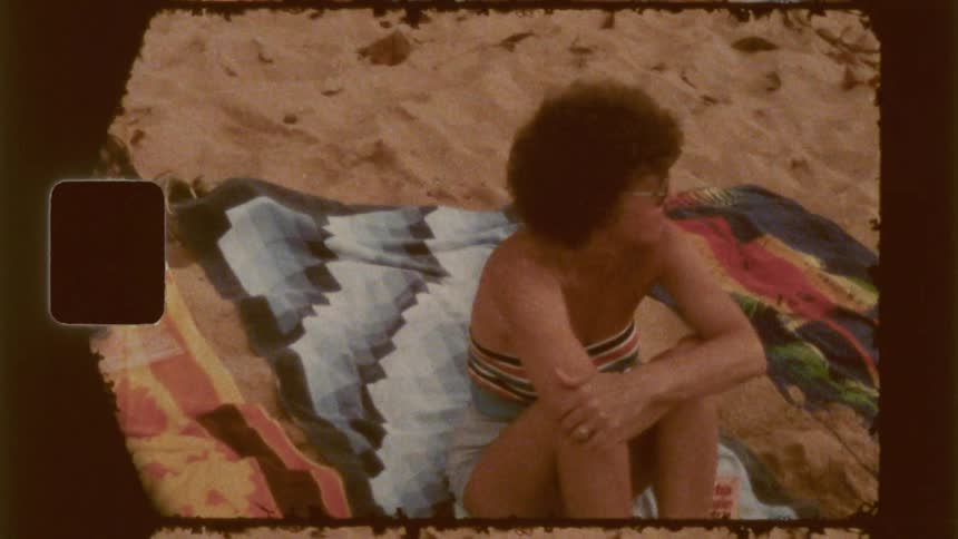 A woman with dark curly hair sitting on a patterned towel on a sandy beach with other beach items nearby