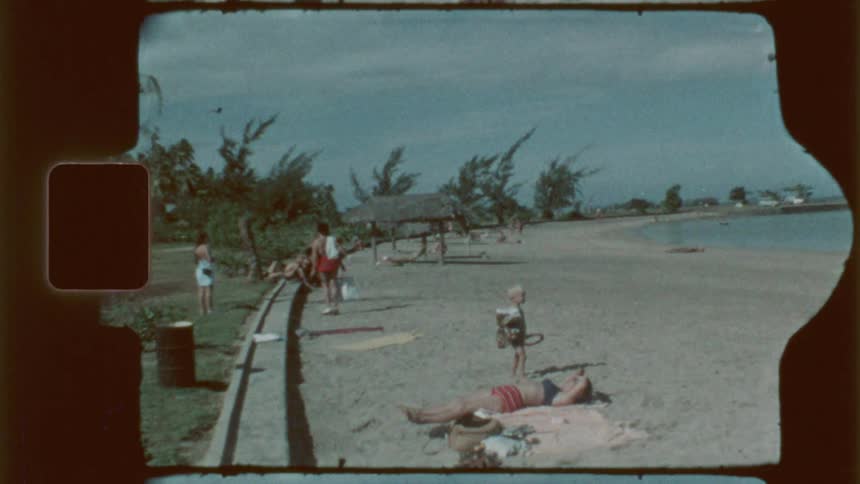 A grainy film frame captures beach scene with several people including a person lying on a towel a child holding a toy