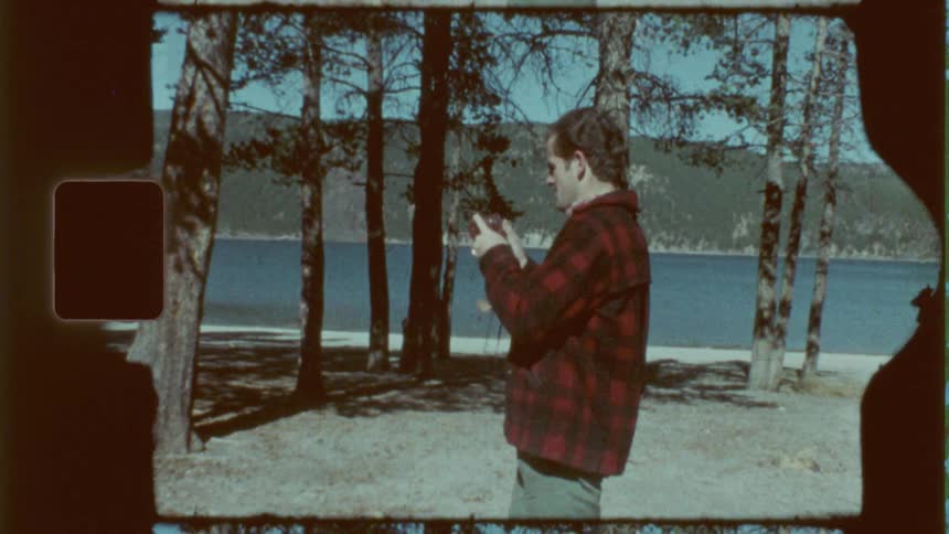 A man with a plaid jacket using a 35mm camera to take pictures there is a lake in the background with trees and mountains which may be what he is taking a picture of