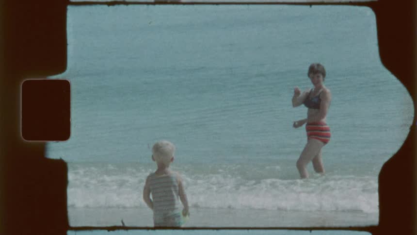 A mother dressed in a vintage swimsuit gestures warmly to her child encouraging them to step into the shallow waters along a serene beach shoreline