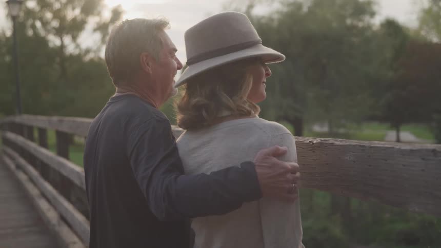 A mature couple stands affectionately on a wooden bridge the mans arm around the womans shoulders as they gaze out at the serene park landscape bathed in soft golden light