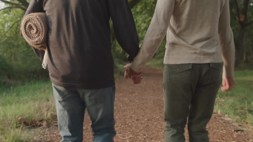 Two people walk hand in hand down a path in a wooded area with one carrying a rolled up mat