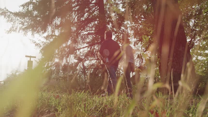 Two people walk together through a grassy area with trees their backs to the viewer in a slightly hazy sunlit setting