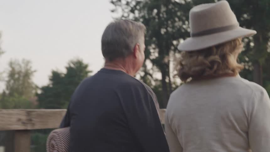 An older couple seen from the back walks together across a wooden bridge surrounded by trees