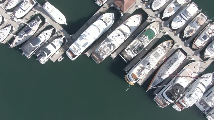 A slow aerial perspective captures numerous white yachts and boats neatly docked in the calm waters of a marina with people walking between platforms