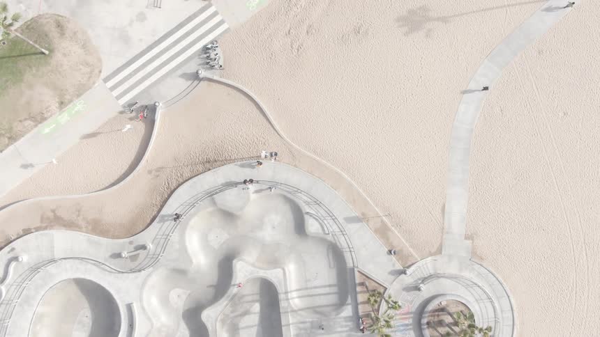 An aerial view shows a concrete skatepark with curved bowls and pathways adjacent to a sandy beach