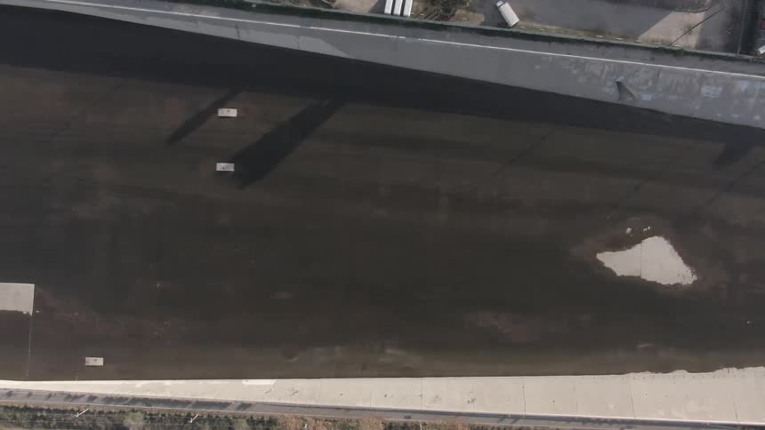An aerial view of a large concrete canal bordered by industrial buildings with small structures casting long shadows and patches of greenery nearby