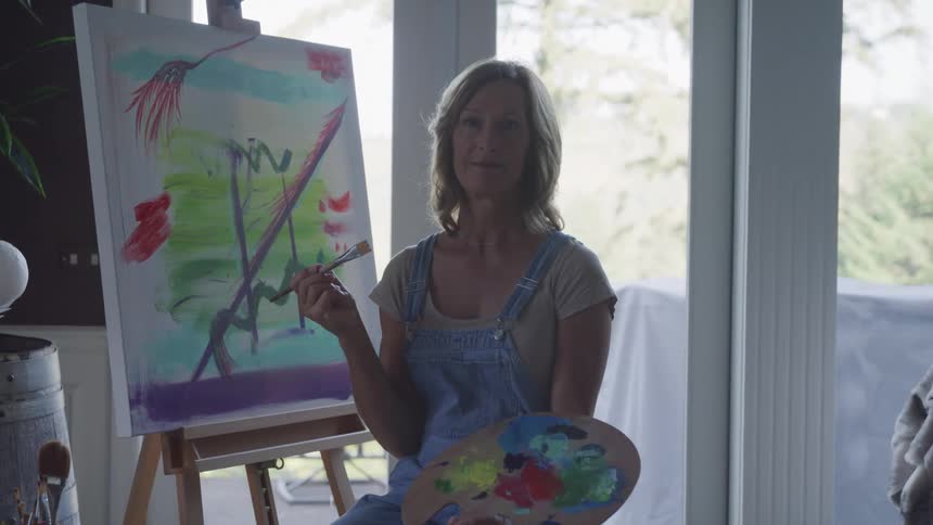 A person stands in front of an easel holding a palette of colors working on a vibrant abstract artwork with green purple and red hues