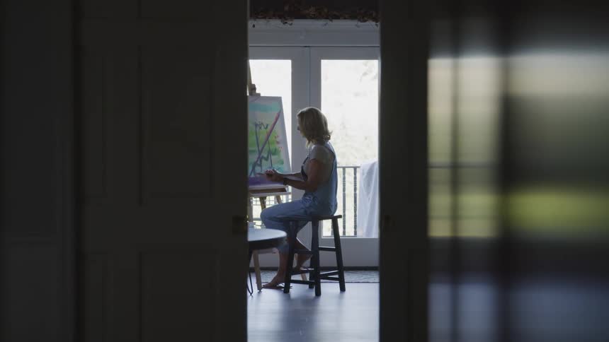 Blonde woman sitting on a stool paints while looking out the glass doors of her house