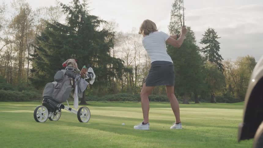 A woman in a skirt and visor swings a golf club on a green course with her golf bag and cart nearby