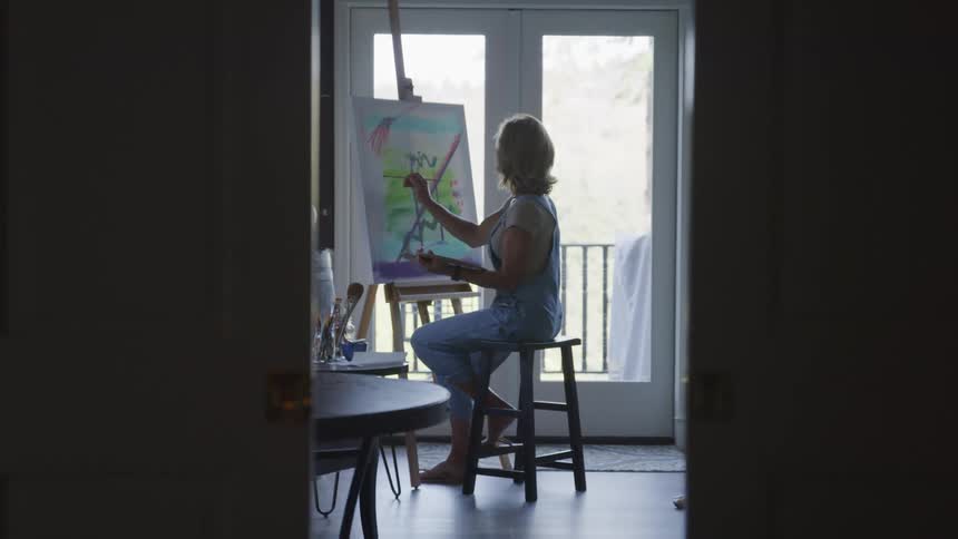 A painter sits on a stool painting at an easel in a room with a brightly lit doorway leading outside