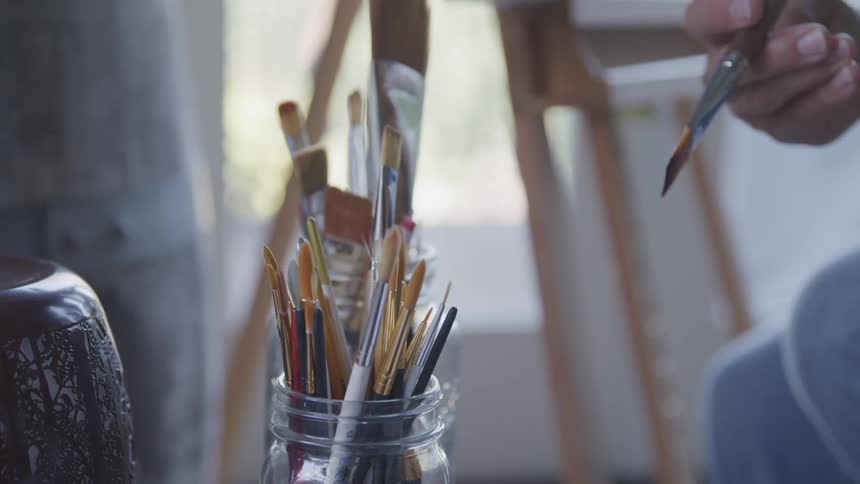 Close up on a glass jar filled with paint brushes as an older mans hand selects one dips it in water offscreen and prepares to paint