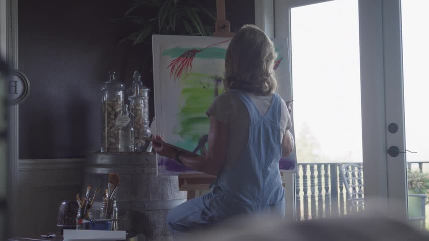 A female painter with blonde hair wearing denim overalls is seen from behind as she paints on a canvas propped on an easel in a bright sunlit room