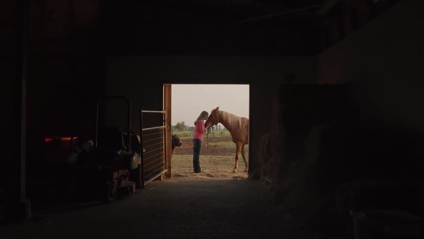 A woman stands in the brightly lit doorway of a dark barn gently kissing a horses forehead