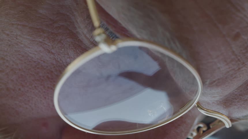 A closeup of a face with blonde eyebrows and golden glasses reflecting light from a table while drawing on paper