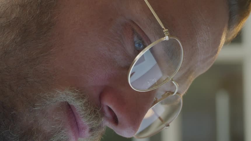 A closeup of a mans face with a beard and glasses illuminated by light reflecting from a screen as he looks attentively