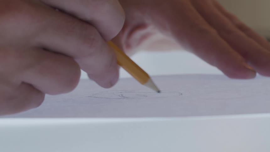 A closeup of a hand drawing on paper with a pencil illuminated by light emanating from the table surface