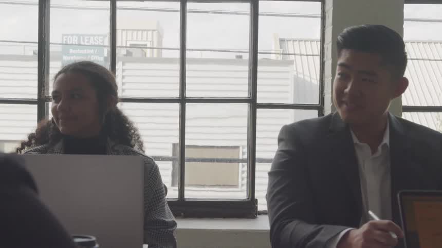 Three individuals are in a professional setting with one standing and gesturing while the other two focus on their laptops at a table