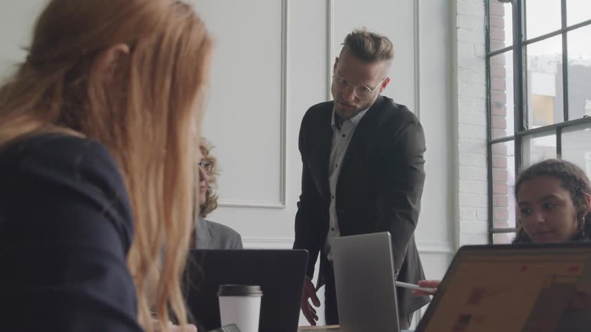A man in a suit stands beside a table where three women are working on laptops in an office setting