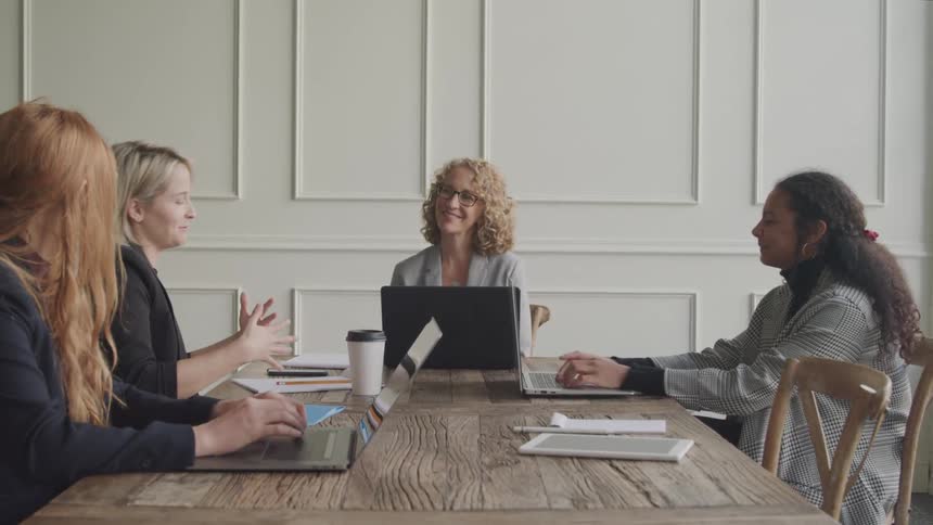 Four businesswomen are gathered around a wooden table in a meeting working on laptops and engaging in conversation