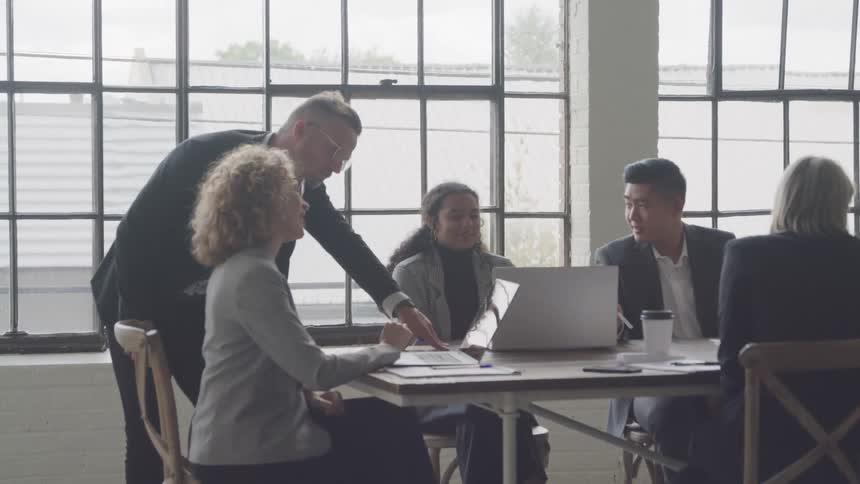 The group of office workers is sitting around the table working and talking except for the man with the glasses he walks to the end of the table to work with the blonde woman in the glasses