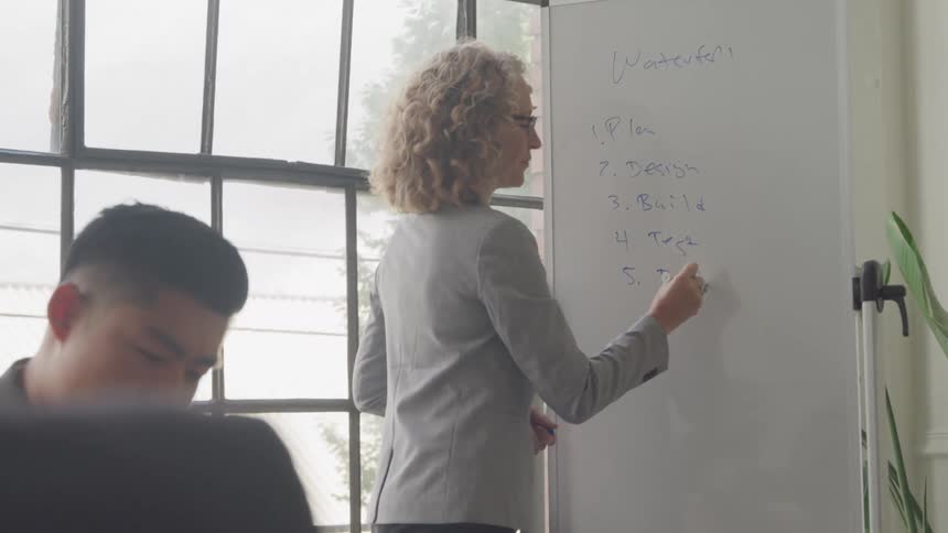 A woman in a blazer writes on a whiteboard listing the steps of a waterfall process in front of an audience in a brightly lit room