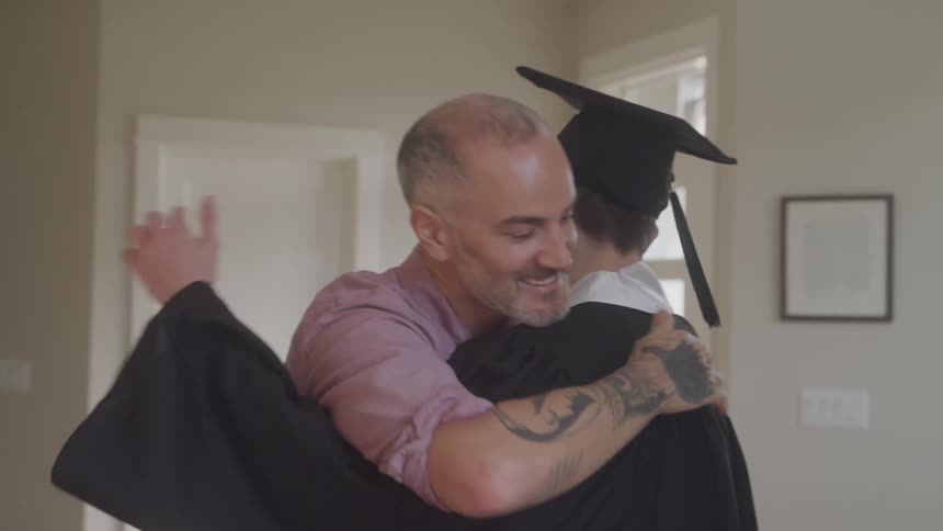 Father adjusting his sons graduation gown and then hugging him contentedly