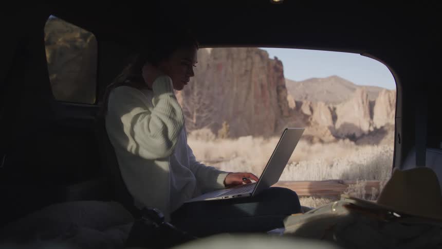 Woman types on a laptop while sitting in the back of a camper in front of mountains