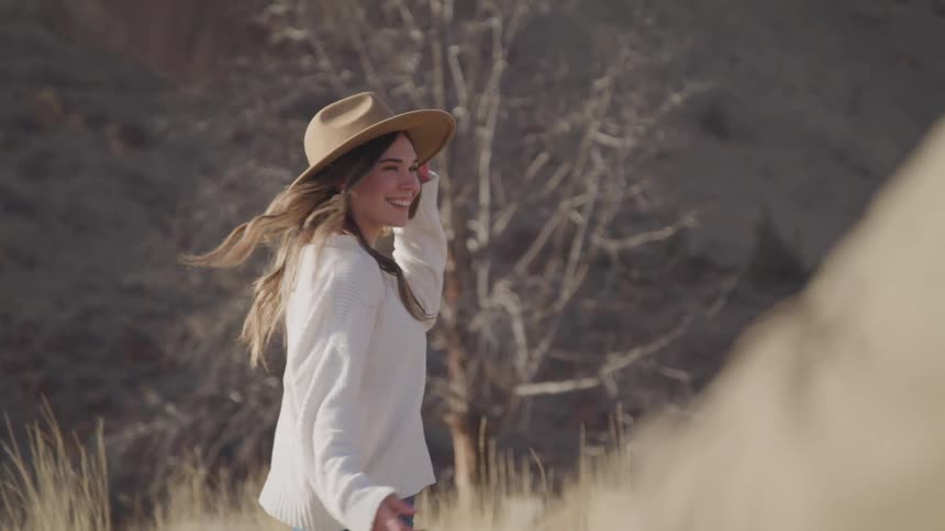A woman in a white sweater and sun hat spins with a smile her long hair flowing surrounded by dry grass a bare tree and a mountain in the background