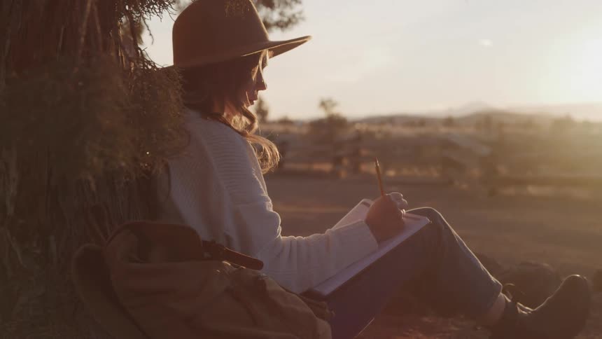 A woman is sitting against a tree and writing in her notebook