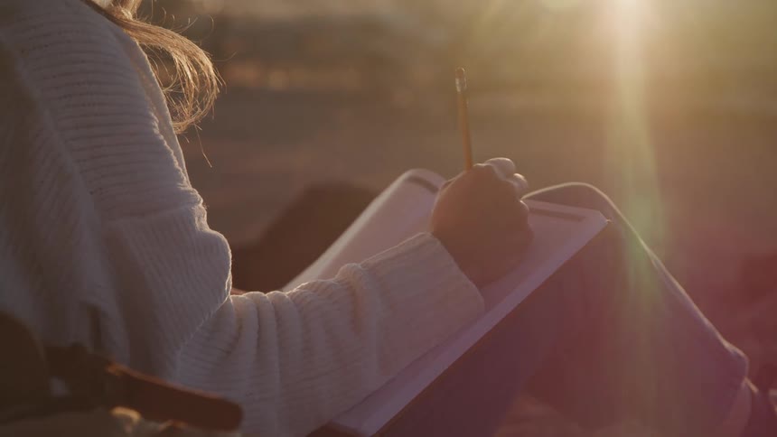 A woman in a white sweater sits with a notebook on her lap writing with a pencil as the warm sunlight bathes her in a golden glow