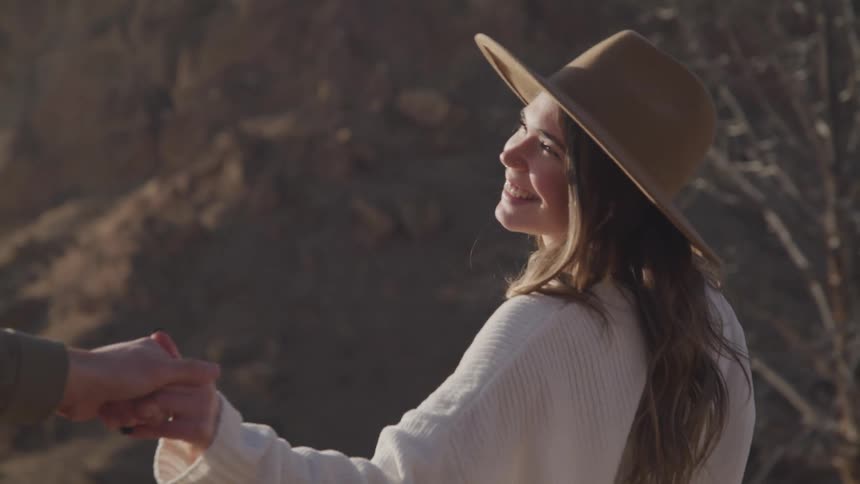 A couple in a desert with mountains in the background as the woman wearing a white sweater and sun hat smiles while posing and holding hands with the man taking her picture