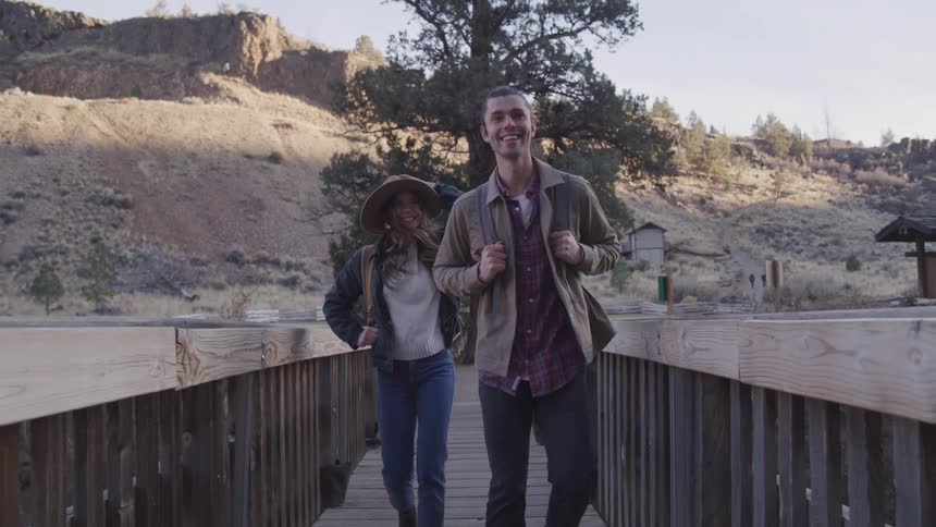 A couple are hiking over a wooden bridge by the mountain