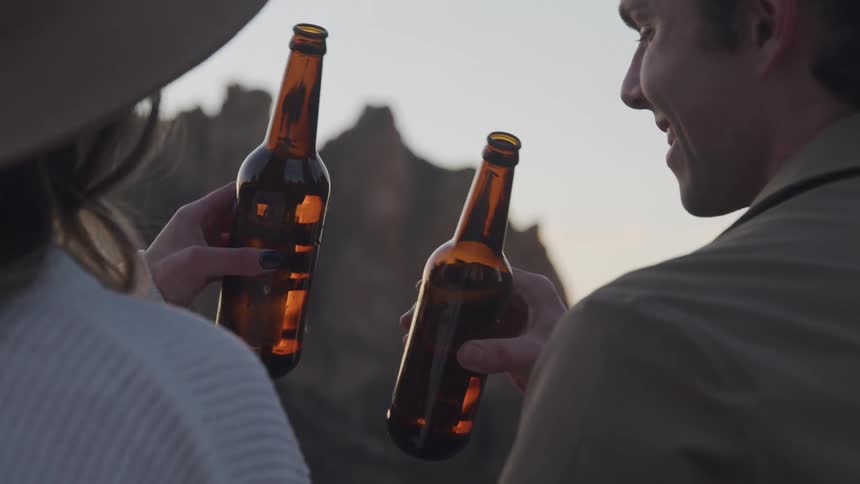 Man and woman clink beer bottles together and drink while gazing at the mountain