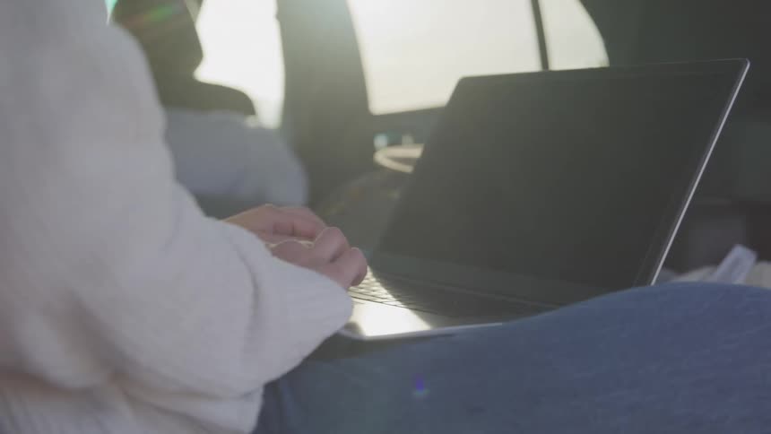 A woman sits in a vehicle working on a laptop with sunlight streaming through the window