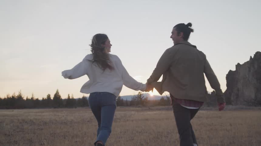 Happy couple running and laughing across a field towards a forest