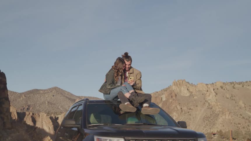 A young couple sits on the roof of a car looking at a phone surrounded by a desert landscape in daytime