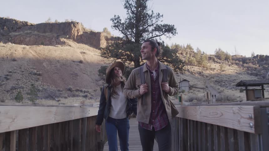 Two people walk on a wooden bridge surrounded by rocky terrain sparse trees and rugged mountains