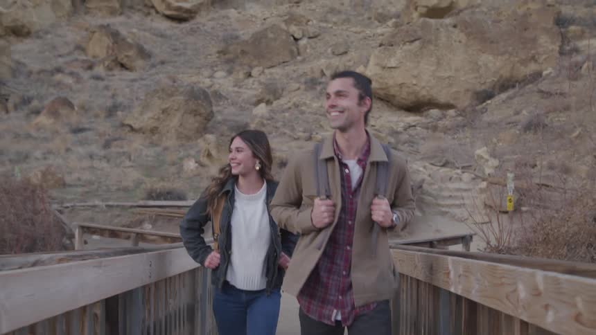 Two people walk along a wooden bridge with rugged rocky terrain and scattered vegetation in the background