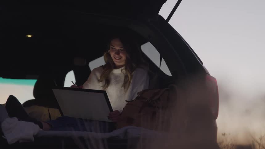 A woman is writing on her tablet while she camps in the hatch of her car