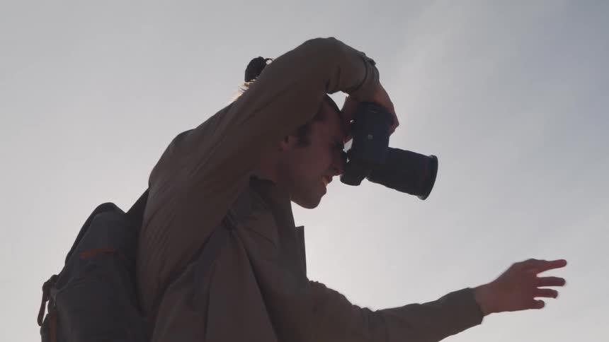 A photographer wearing a brown shirt and a black backpack holding a camera up to his eye to take a photograph