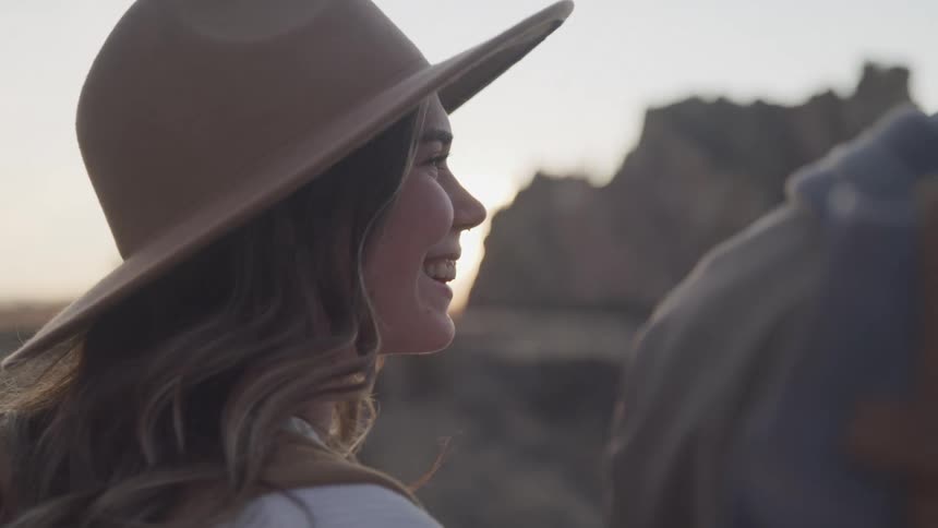 A woman with wavy hair wearing a hat standing outside against a backdrop of rock formations at dusk