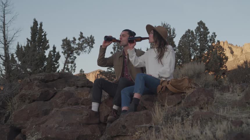 Man and woman clink their bottles while sitting on boulders in the wilderness near the mountains