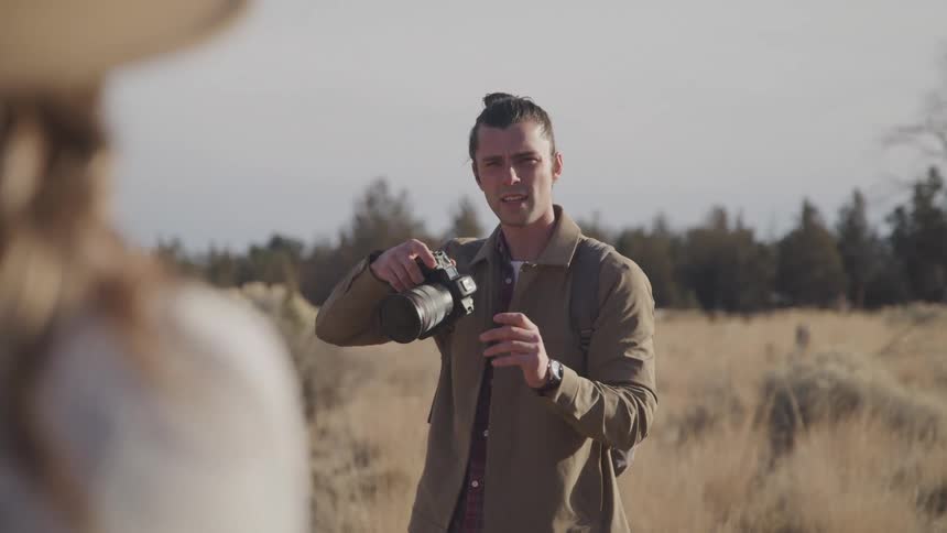 A photographer is pointing the camera at a woman modeling in a dry grass field on a sunny fall day