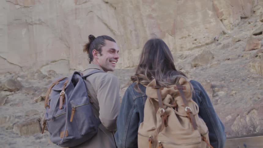 Two people carrying backpacks walk on a wooden bridge surrounded by rocky terrain and rugged mountains
