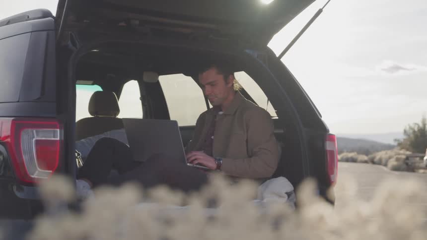 A man sits in the open trunk of an suv typing on a laptop with a smile surrounded by sunlight in an outdoor setting