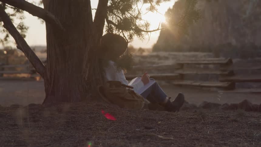 A woman wearing a hat is sitting under a tree on a farm writing in a notebook