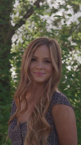 A young woman with long wavy blonde hair smiles gently at the camera with a backdrop of green foliage