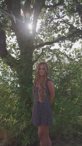 A young woman with long wavy blonde hair stands in a natural setting wearing a short dress with a large tree and foliage behind her and sunlight filtering through the leaves